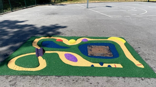 Circuit de billes en sol souple vert dans la cour ensoleillée de l'école des Fifendes, avec sa piste jaune et sa plaque de fonte entourée d'un bassin bleu