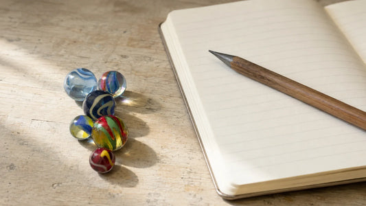 Sept billes en verre colorées posées en petit tas sur une table de bois clair, à côté d'un cahier d'écolier ouvert aux lignes vides et d'un crayon de bois posé en travers