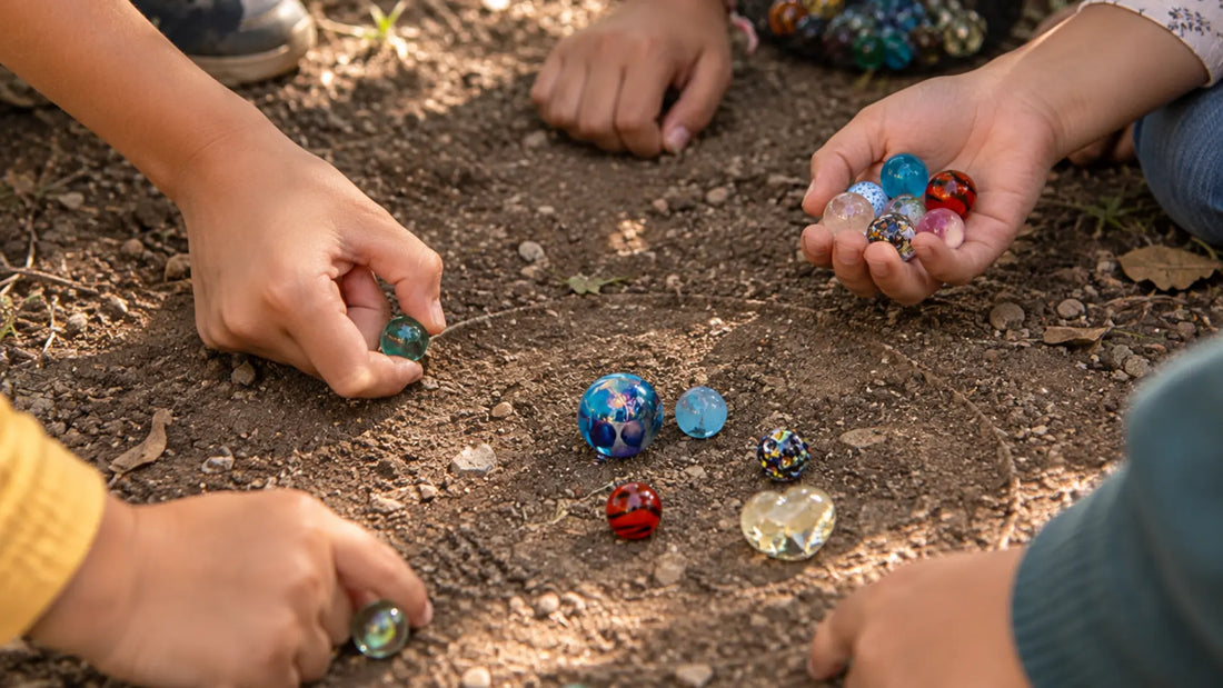 Plusieurs mains d'enfants réunies autour d'un cercle tracé dans la terre d'une cour, billes de verre MesBilles posées au sol et une poignée de billes tenue dans une paume ouverte