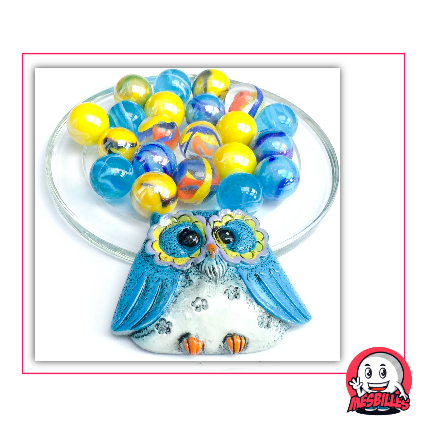 Hibou bleu en céramique accompagné de 20 billes de 16 mm en verre bleu, jaune, transparent et marbré, posées sur une coupelle en verre avec encadrement rose et logo MesBilles.